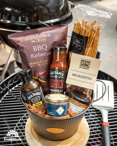 Panier garni cadeau pour barbecue avec sauces, chips et snacks, posé sur un gril.